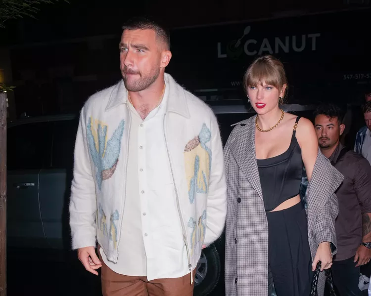 Taylor Swift and Travis Kelce seen leaving SNL. Swift is seen wearing a black corset top that featured gold-chain detailing on the straps with a pair of loose-fitting trousers and an over-sized gray plaid trench coat. Her blonde hair was pulled back in a ponytail and she sported her signature red lip. Kelce is seen wearing brown leather pants, a white button-down layered underneath a cream-colored embroidered jacket, and white sneakers. 